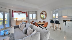 Una sala moderna con un sofá gris, una mesa de comedor y sillas. Hay una zona de cocina con armarios blancos y un ventanal grande.