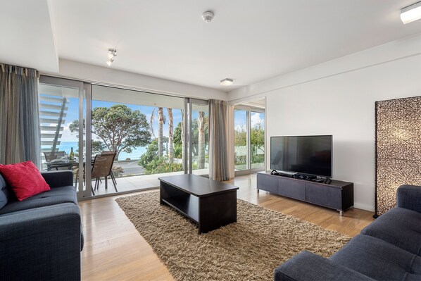 Una sala de estar moderna con un televisor grande, sofás oscuros, una mesa de centro y vistas al exterior.