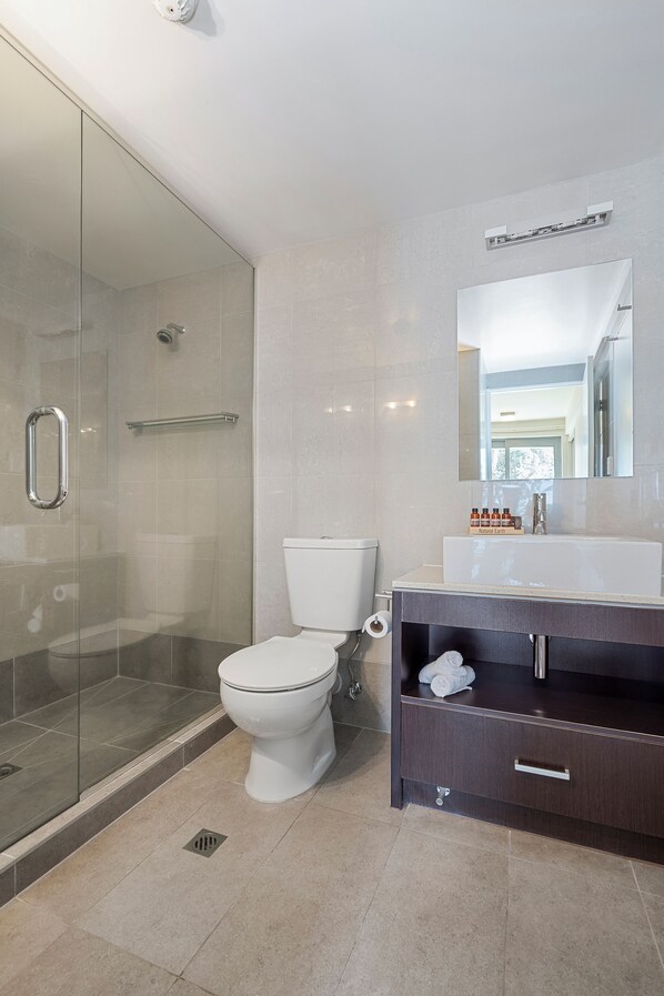 Un baño moderno con cabina de ducha de vidrio, inodoro blanco, tocador con mueble oscuro y un espejo grande sobre el lavabo.