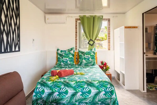 A bedroom with a bed, a window with green curtains, a white air conditioning unit, and a brown chair. - Nahi Loge, Welcome home (Vaitape)