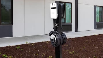 En elbil-ladestation med et kabel viklet om en stang, beliggende i et anlagt område med barkflis og små planter.