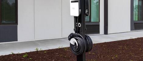 Una estación de carga para vehículos eléctricos con un cable enrollado alrededor de un poste, ubicada en un área ajardinada con mantillo y plantas pequeñas.