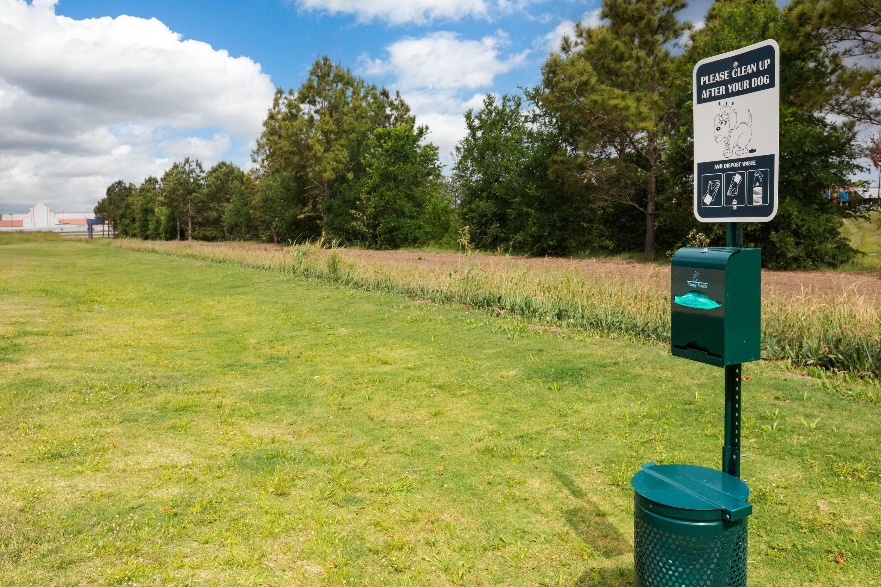 A grassy field with a sign instructing to clean up after dogs and dispose of waste, a green trash bin, and a Poopy Pouch.