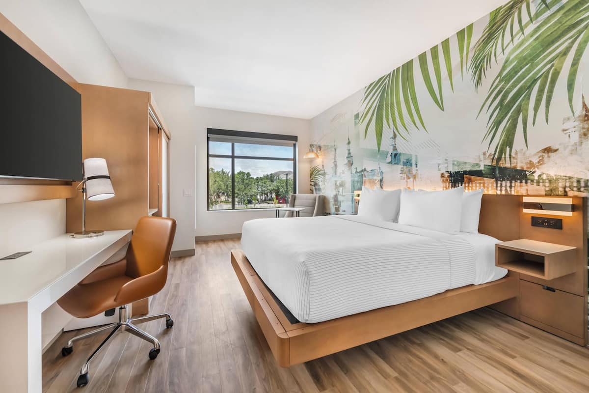 Une chambre d’hôtel moderne, dotée d’un grand lit, d’un bureau et d’une chaise. Une fenêtre offre une vue sur la verdure environnante.