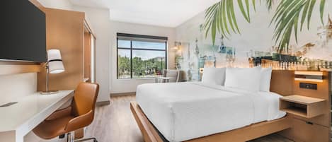 A modern hotel room with a large bed, a desk, and a chair. There is a window with a view of greenery outside.