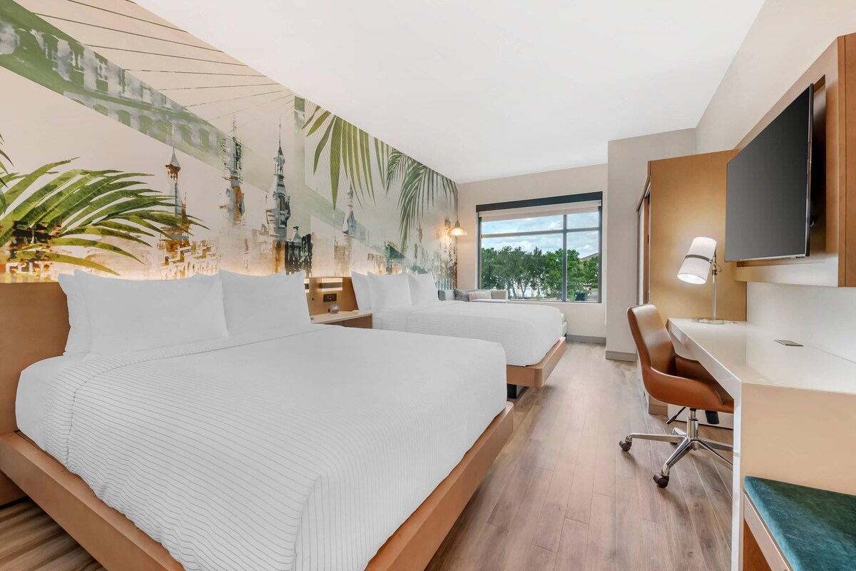 Ein modernes Hotelzimmer mit einem großen Bett, einem Schreibtisch und einem Stuhl. Ein Fernseher ist an der Wand befestigt und ein Fenster bietet Blick ins Freie.