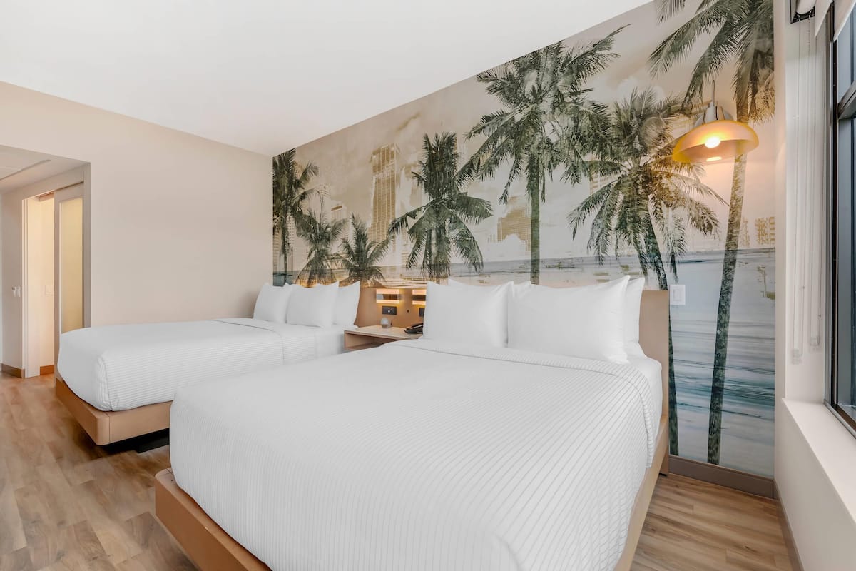 Une chambre d’hôtel avec deux lits, une grande fresque représentant une plage sur le mur et une fenêtre donnant sur des palmiers.
