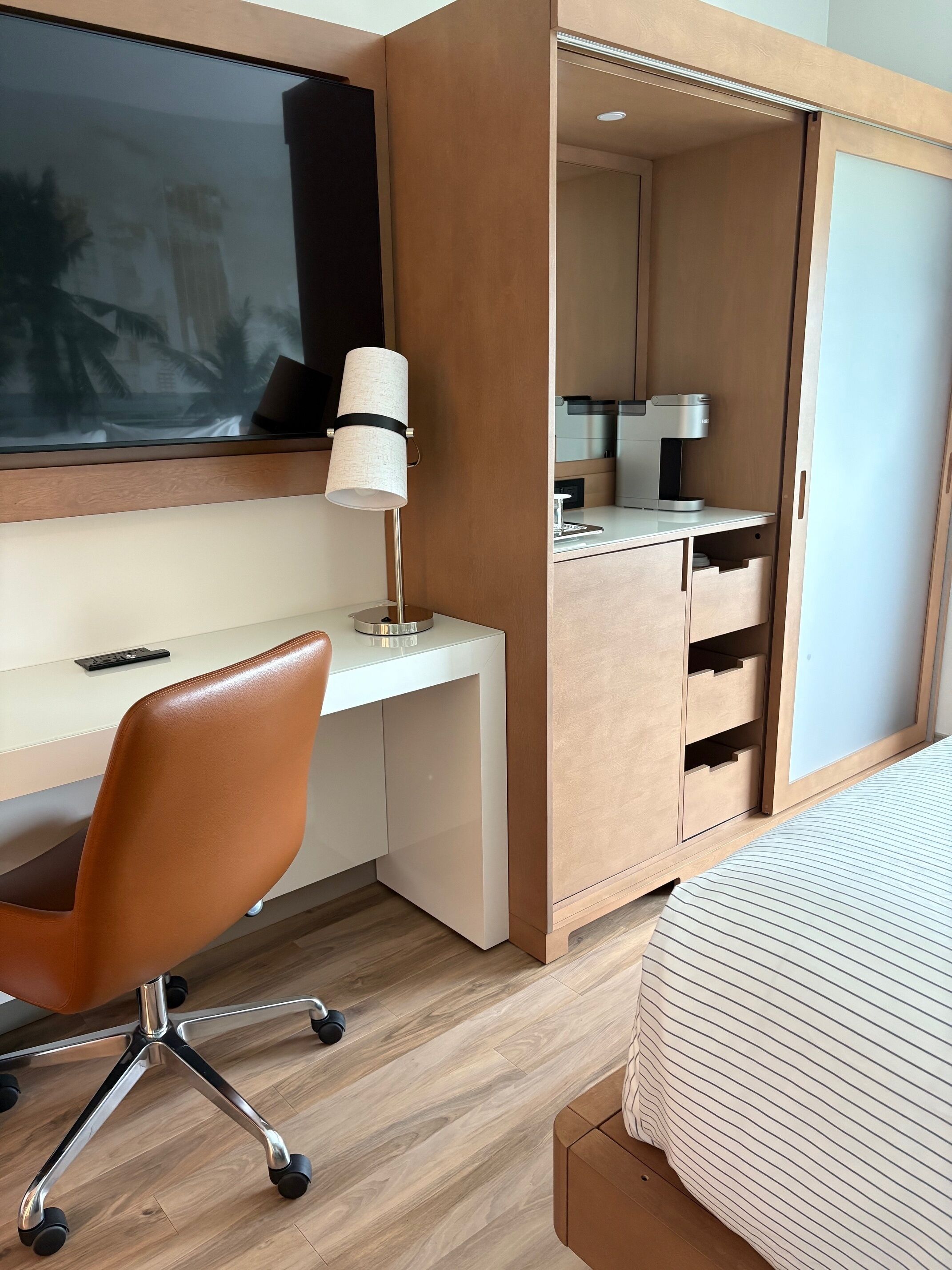 A modern hotel room with a wooden desk, a swivel office chair, a bedside table, a bed with striped bedding, a large window, and a built-in wardrobe.
