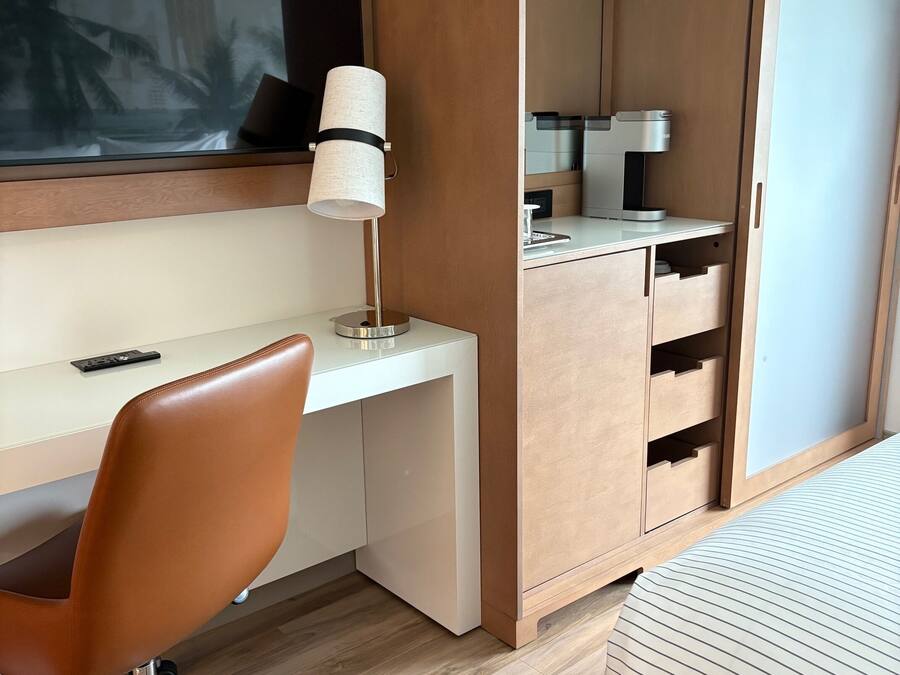 Et moderne hotelværelse med skrivebord i træ, en drejelig kontorstol, et natbord, seng med stribet sengetøj, et stort vindue og en indbygget garderobe.
