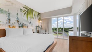 A modern hotel room with a large bed, a desk, a chair, and a large window offering a view of the city. - Cambria Hotel Tampa-Brandon (Tampa)