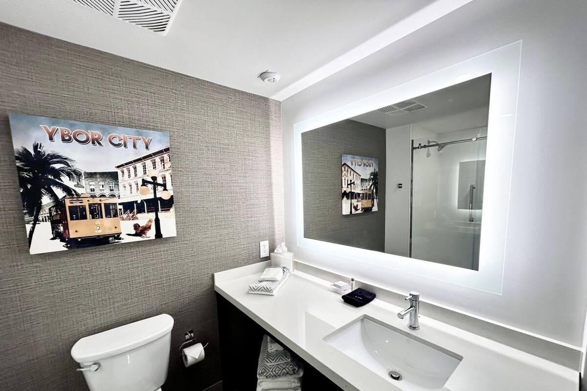 Ein modernes Badezimmer mit einem großen Spiegel, einem weißen Waschbecken und einer Toilette. An der Wand hängt ein gerahmtes Bild einer Stadtansicht.