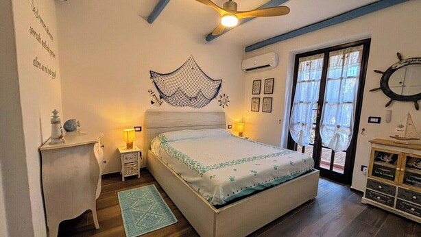 A bedroom with a bed, bedside lamps, a ceiling fan, a window with curtains, a wall clock, and a small table with a lamp.