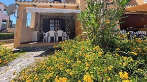 A building with a stone facade, a covered patio with a table and chairs, and a garden with yellow flowers.