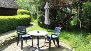 A patio with a round table and four chairs, surrounded by a stone-tiled floor and lush greenery. - Ferienwohnung im Reetdachhaus, Burhave (Burhave)