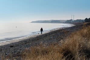 Eine Küstenlandschaft mit kiesigem Strand, einer Person, die spazieren geht, und Windkraftanlagen in der Ferne.