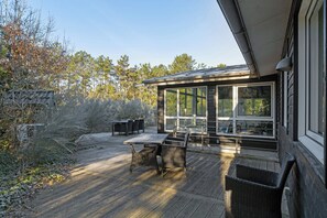 A covered outdoor patio with a table and chairs, a glass-enclosed room, and a view of trees.