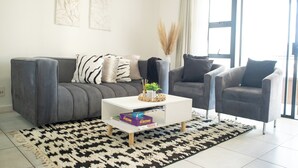 Una sala de estar moderna con un sofá gris, una mesa de centro blanca y un sillón gris. Hay dos cuadros en la pared, un jarrón con plantas secas y una alfombra estampada en el suelo.