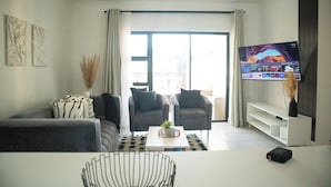 Una sala moderna con un sofá, dos sillones, una mesa de centro con una planta, un televisor de pantalla plana y un amplio ventanal con cortinas.