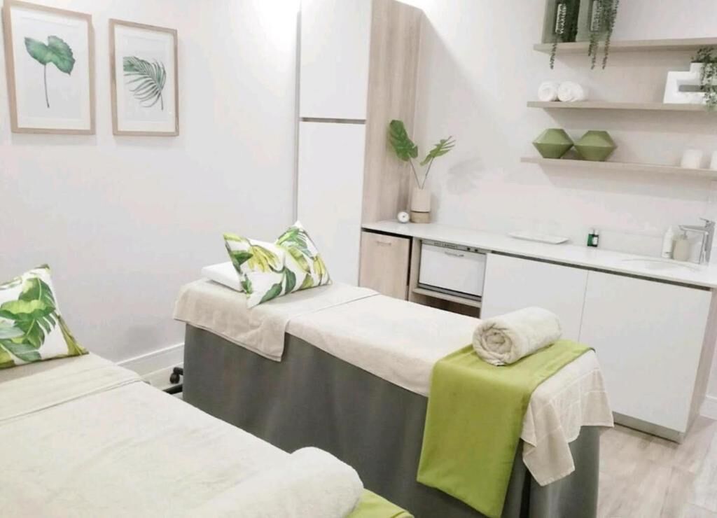 A spa room with two massage tables, a small sink, and shelves with plants and toiletries.