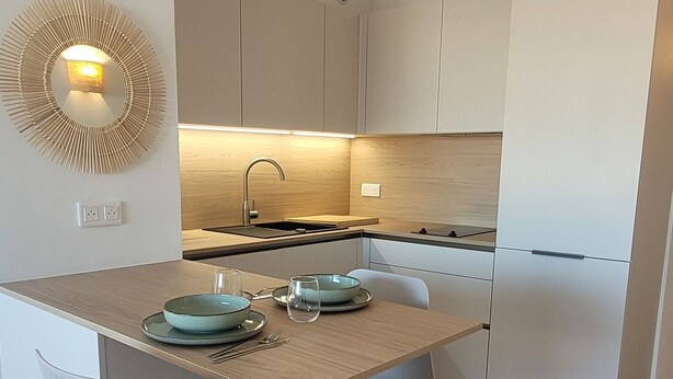 A modern kitchen with white cabinetry, a wooden countertop, and a dining area with a white chair and table. - Cosy apartment large T1 of 32m2, direct access to the beach (Mauguio)