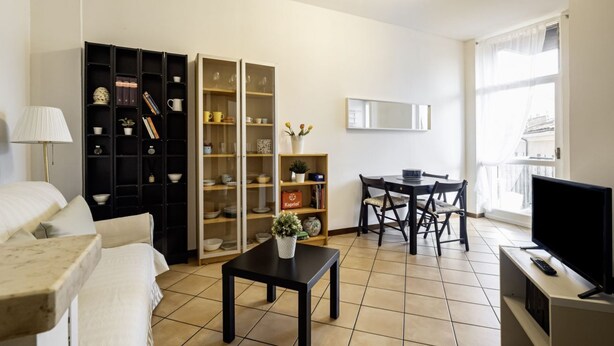A living room with a sofa, a coffee table, a TV, a dining table with chairs, and two shelving units with various items. - GuestHost - Naviglio Pavese Comfortable Apt with Balcony! (Milano)