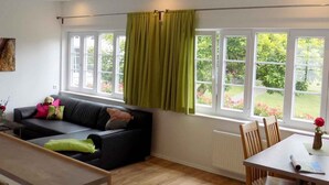 A modern living room with a black sofa, a dining table with chairs, and a view of a garden through large windows.