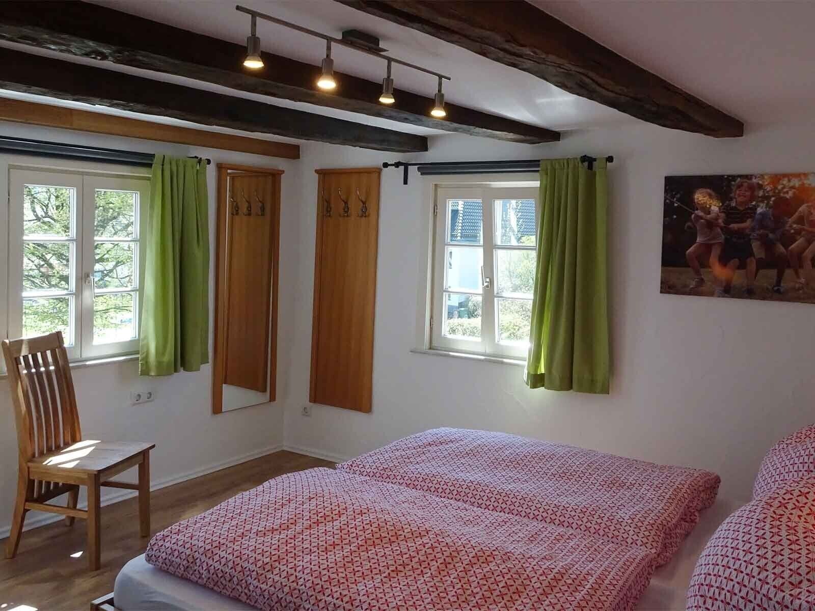Een kamer met een bed, een stoel, een raam met gordijnen, een schilderij aan de muur en een plafond met zichtbare balken.