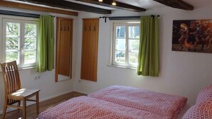 A room with a bed, a chair, a window with curtains, a painting on the wall, and a ceiling with exposed beams.