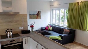 A modern kitchen with a built-in oven, a kettle, and a vase with flowers. There is a black sofa with cushions and a teddy bear. A window with green curtains is visible.