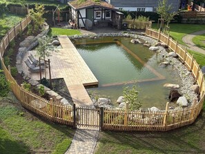 Ein runder Teich mit einer Holzterrasse, umgeben von einem Holenzaun und einem kleinen Gebäude mit geneigtem Dach.