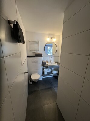 A narrow bathroom with white tiled walls, a white toilet, a sink with a mirror above it, and a towel rack.
