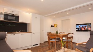 Une cuisine moderne avec des meubles blancs, un four à micro-ondes et un tableau noir. On y trouve un coin repas avec des chaises et une table en bois, ainsi qu’un téléviseur fixé au mur.