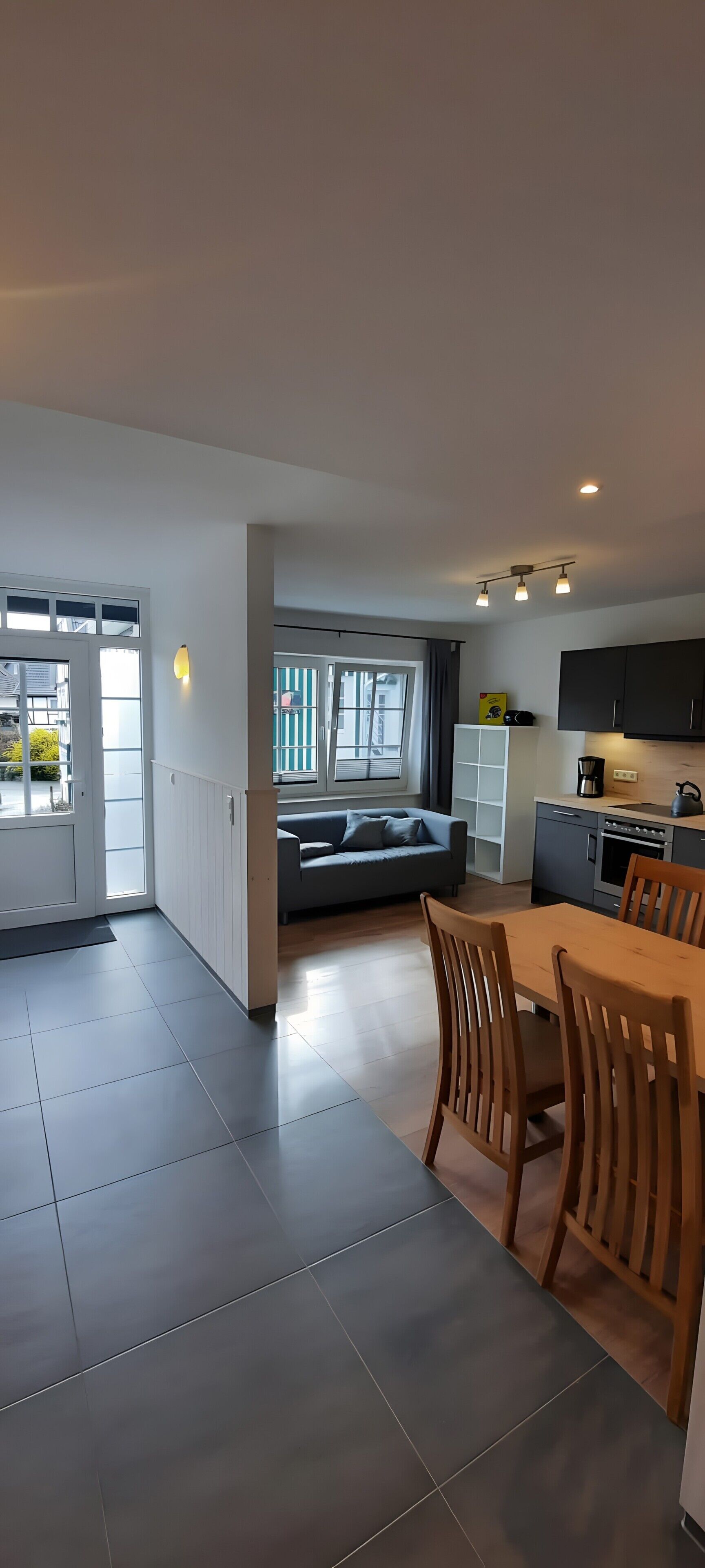 A modern kitchen with a dining area, a sofa, and a view through a glass door.