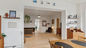Un espacio amplio con piso de madera, una mesa de comedor con sillas, un sofá y una lámpara. Hay estantes con objetos decorativos y un cuadro en la pared.