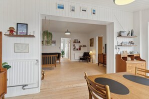 A spacious room with a wooden floor, a dining table with chairs, a sofa, and a lamp. There are shelves with decorative items and a framed picture on the wall.