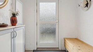 Una puerta blanca con un panel de vidrio esmerilado, un banco de madera, un florero con flores y un reloj fijado a la pared.
