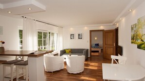 A modern living room with a sofa, two white chairs, a dining table, and a kitchen area with a window. - Apartment 'Sonnenblume' with Private Terrace, Shared Garden and Wi-Fi (Finnentrop)