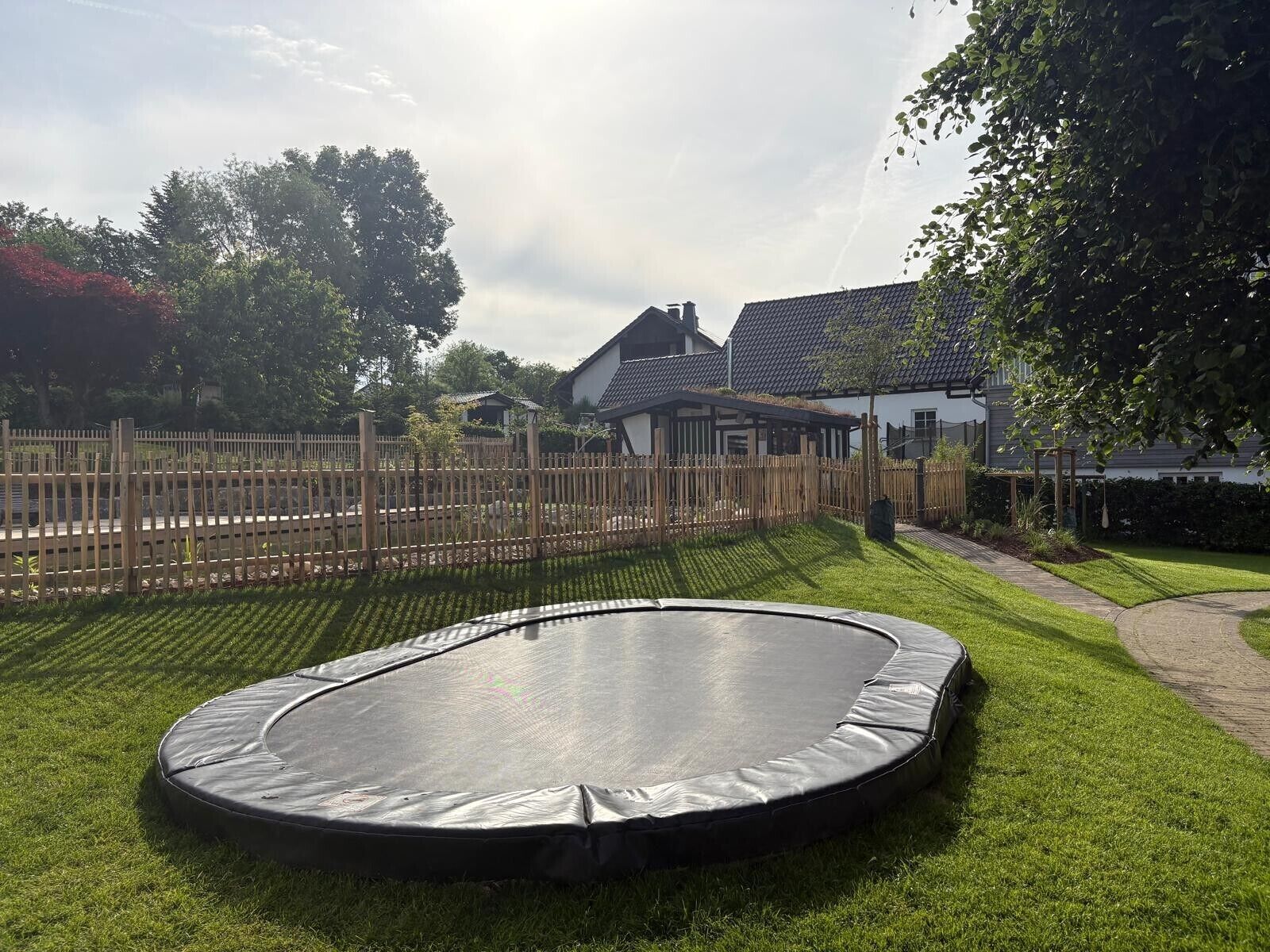 Un trampolino in un giardino privato con recinzione in legno, un prato e una casa sullo sfondo.
