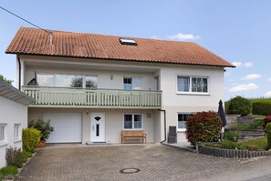 Une maison à deux étages, avec un toit en tuiles, un porche à l’avant, un banc et un petit jardin.
