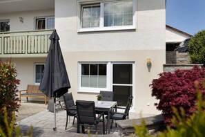 Un bâtiment à deux étages doté d’un balcon, d’une terrasse avec une table et des chaises, et d’un arbuste aux feuilles rouges.