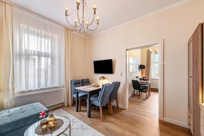 A modern living room with a dining area, a TV mounted on the wall, and a large window with sheer curtains. - Holiday Apartment 'Seestern' with Shared Garden and Wi-Fi (Kühlungsborn)