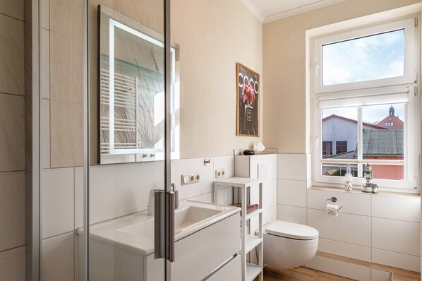 Ein modernes Badezimmer mit Duschkabine aus Glas, Toilette, Waschbecken und einem Fenster mit Blick auf Gebäude.