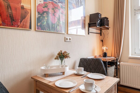 Un comedor con una mesa de madera, platos blancos, tazas y un jarrón con flores. Hay dos cuadros en la pared, un altavoz negro sobre una estantería y una ventana con cortinas.