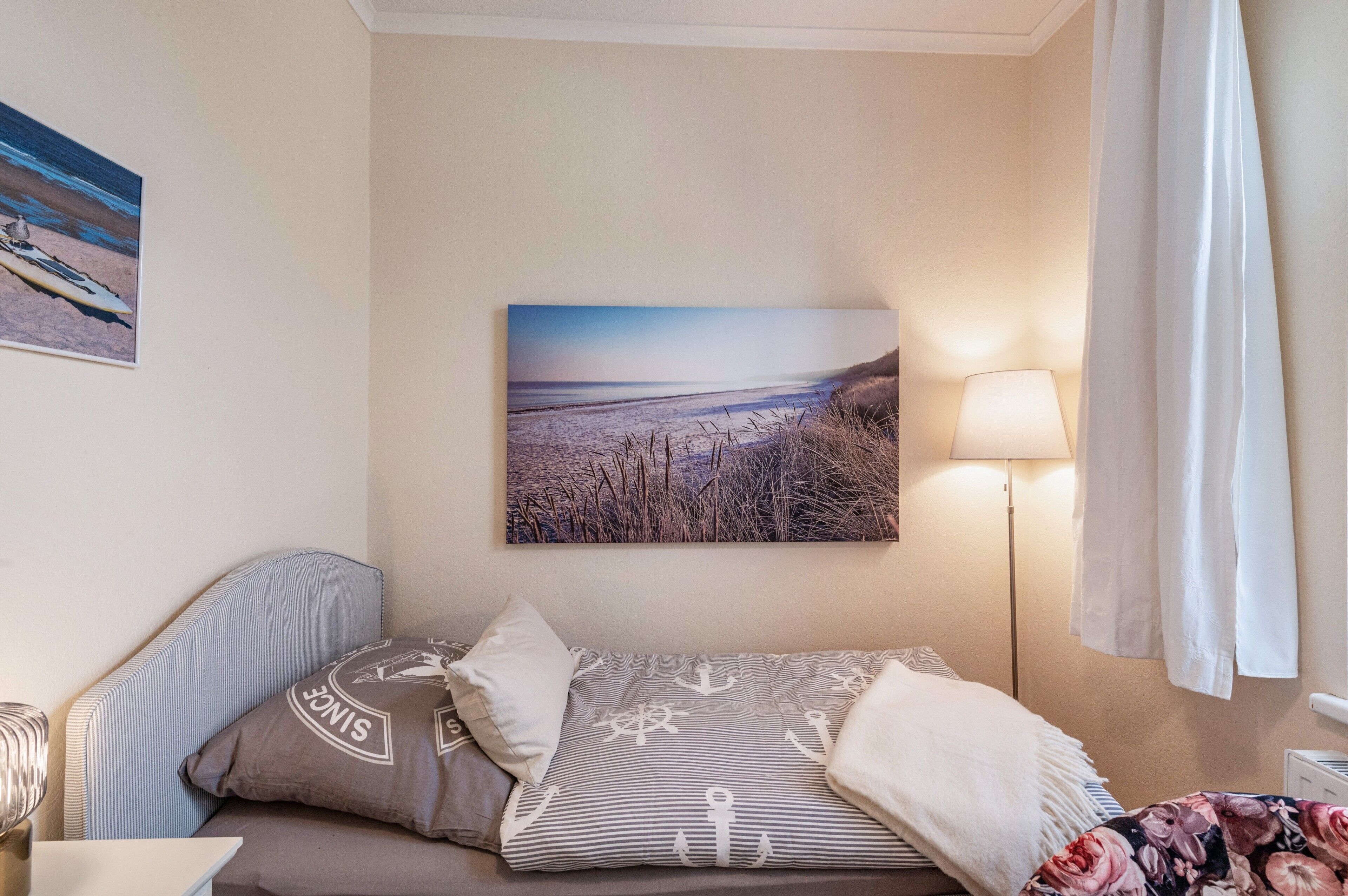 A room with a bed, a wall-mounted lamp, a painting, and a window with curtains.