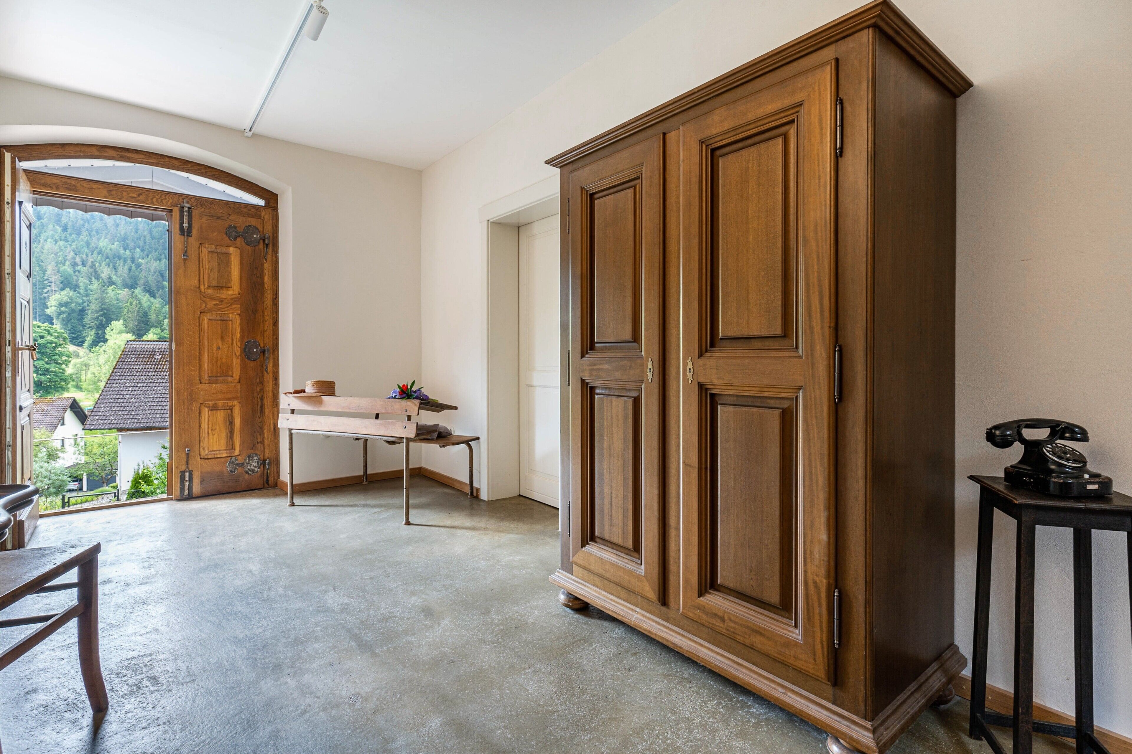 Une pièce avec une grande armoire en bois, une petite table avec une lampe et une vue sur l’extérieur par une porte ouverte.