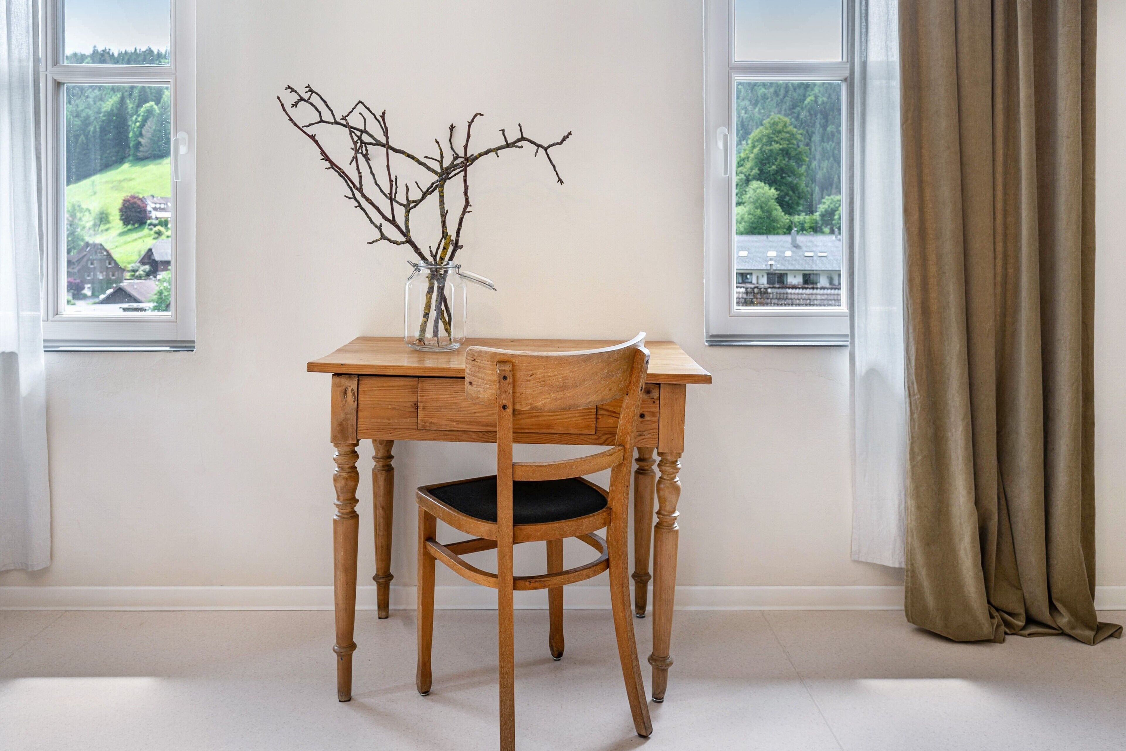 Een houten tafel en stoel in een kamer met twee ramen en uitzicht op een groen landschap.