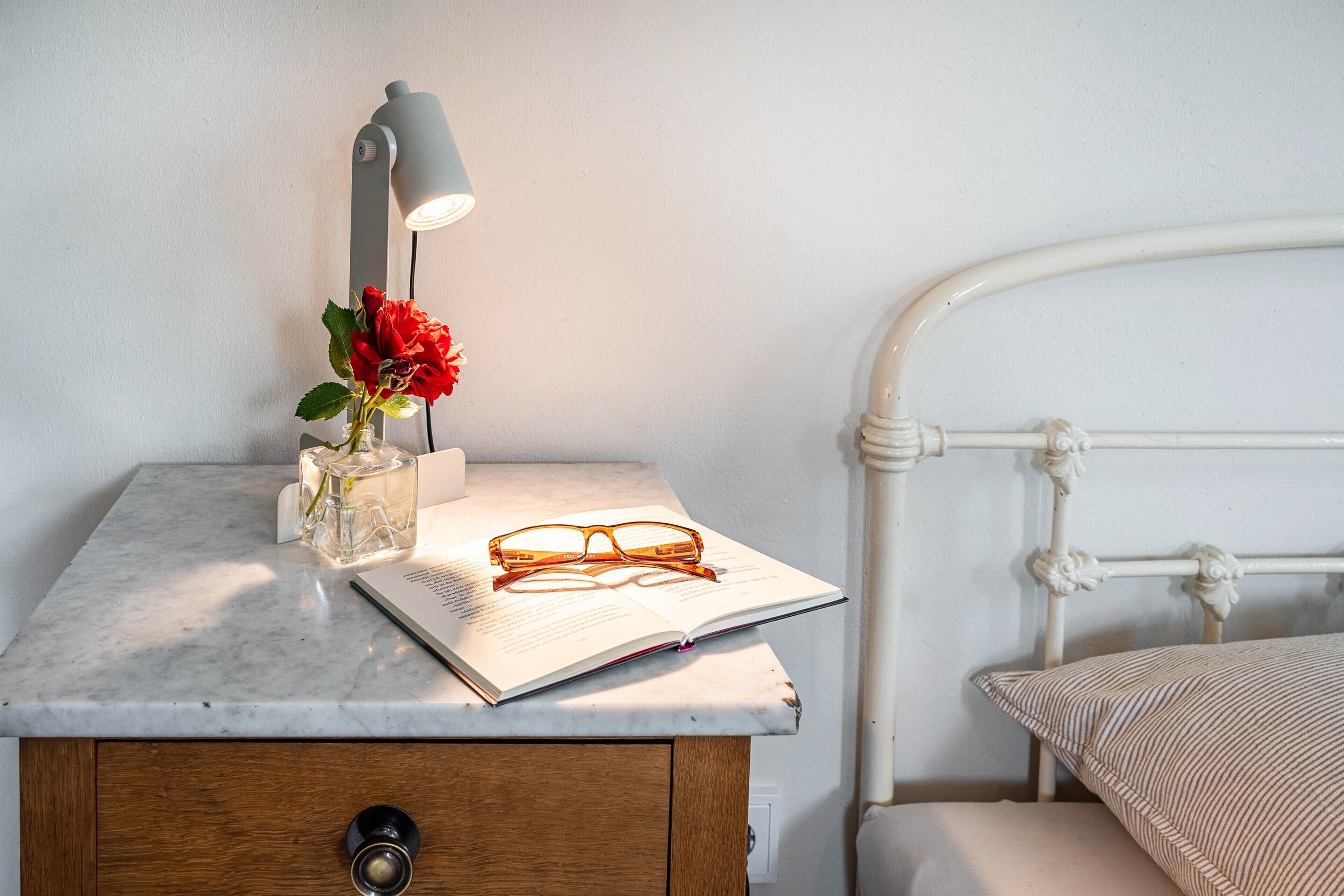 A bed with a white metal frame, a bedside table with a lamp, a book, and a pair of glasses.