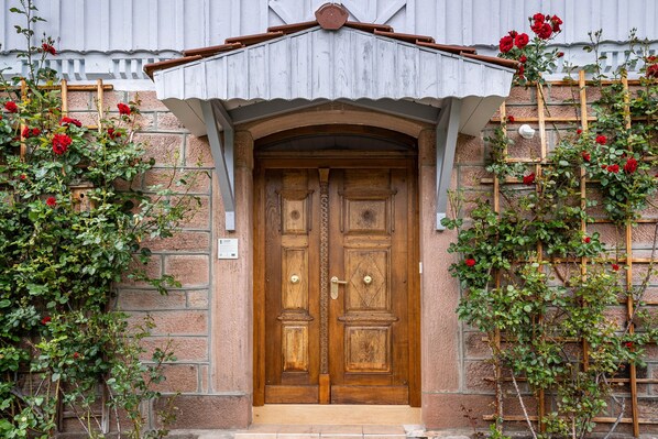 A wooden double door with a decorative arch above it, flanked by climbing roses and a wooden trellis.