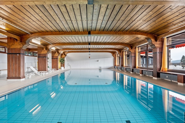 Une grande piscine intérieure avec des poutres en bois, des carreaux bleu clair et des chaises longues disposées le long des bords.
