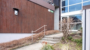 Un bâtiment doté d’une façade en bois, d’un escalier menant à une porte et d’un petit jardin agrémenté de plantes et d’un arbre.